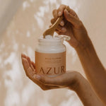 Close-up van handen die met een houten lepel wit magnesium badzout scheppen uit een glazen pot van het merk AZUR Wild Flower.