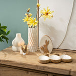 Landelijke paasstyling op een houten dressoir met de konijnen serveerschaal, witte vazen met gele bloemen en een houten paasdecoratie