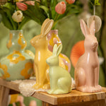 Een sfeervolle opstelling van roze,gele en groene keramische haasjes en vazen op een houten bankje, een modern paasaccent voor het voorjaar.