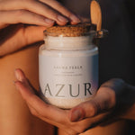 Close-up van een glazen pot AZUR Sauna Feels badzout die met beide handen wordt vastgehouden tijdens zonsondergang, wat een warme en rustgevende sfeer creëert.