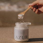 Een glazen pot AZUR Sauna Feels magnesium badzout op het strand, waarbij iemand met een houten lepel wit badzout boven de pot houdt tegen een achtergrond van de zee.