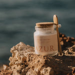 Een glazen pot AZUR Sunset Dreams magnesium badzout rustend op een rotsachtige kustlijn met de blauwe zee op de achtergrond.