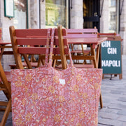 Een roze en oranje gewatteerde tas hangt aan de leuning van een rode houten terrasstoel bij een café.