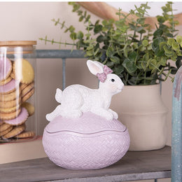 Roze bewaardoosje in de vorm van een paasei met een wit konijntje op een houten plank naast een pot met koekjes