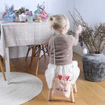 Een kind zit op een krukje met een roze fluwelen konijnentasje op de rug bij een vaas met paastakken