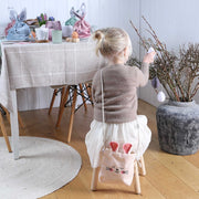 Een kind zit op een krukje met een roze fluwelen konijnentasje op de rug bij een vaas met paastakken