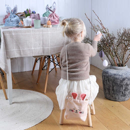 Een kind zit op een krukje met een roze fluwelen konijnentasje op de rug bij een vaas met paastakken