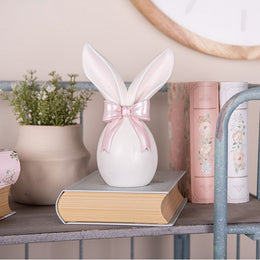 Wit decoratiebeeld van konijnenoren met een roze strik op een stapel boeken in een landelijk interieur