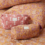 Close-up van een handbedrukte oranje-roze quilted toilettas met een paars kwastje aan de rits, liggend op bijpassend beddengoed.