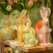Een sfeervolle opstelling van roze,gele en groene keramische haasjes en vazen op een houten bankje, een modern paasaccent voor het voorjaar.