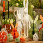 Sfeervolle paastafel met verschillende witte en groene paasaccenten, keramische vazen en papieren paasversiering.