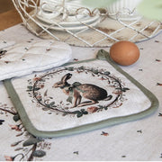 Landelijke beige pannenlap met groene bies en een print van een konijn in een krans, liggend op een bijpassend tafelkleed.