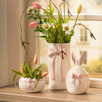 De witte keramische vaas met roze strik gestyled in een vensterbank met witte tulpen en voorjaarstakken, naast de bijpassende paasdecoratie.