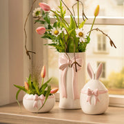 De witte keramische vaas met roze strik gestyled in een vensterbank met witte tulpen en voorjaarstakken, naast de bijpassende paasdecoratie.