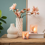 Twee brandende roze windlichten met konijntjes gestyled op een houten tafel naast moderne witte vazen en bloesemtakken.